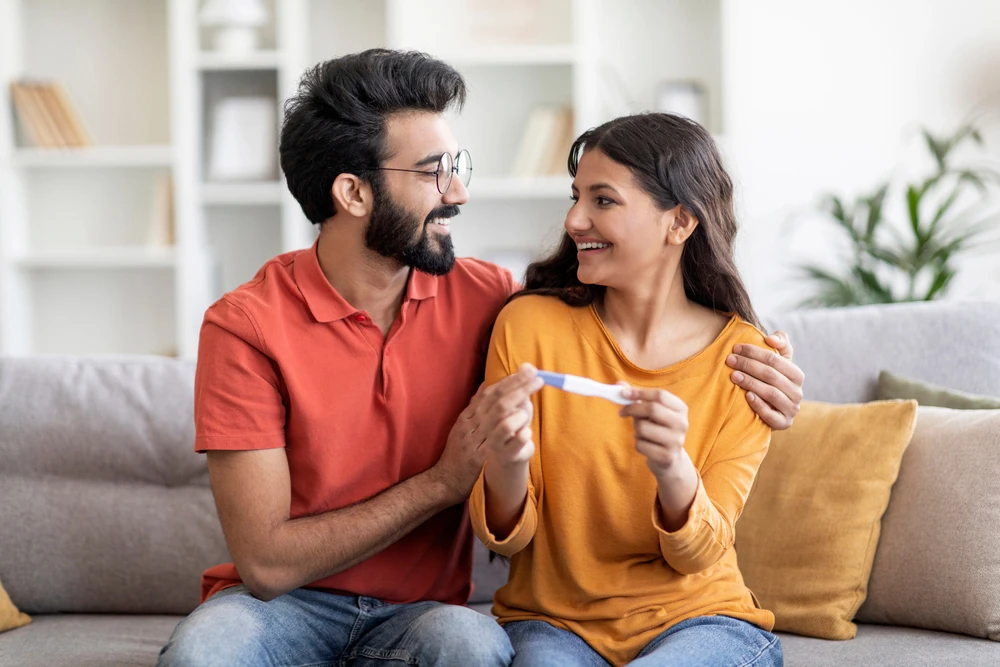 Happy couple holding positive pregnancy test and embracing at home, romantic young eastern spouses waiting for baby, celebrating becoming parents, sitting on couch in living room, closeup